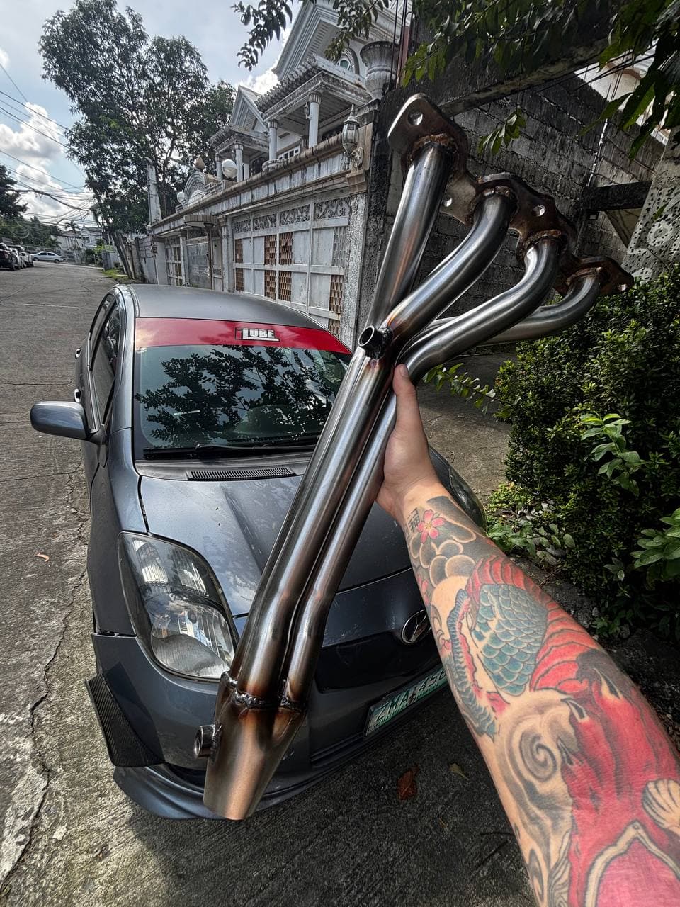 Stainless tubular exhaust header held in front of a gray Toyota
